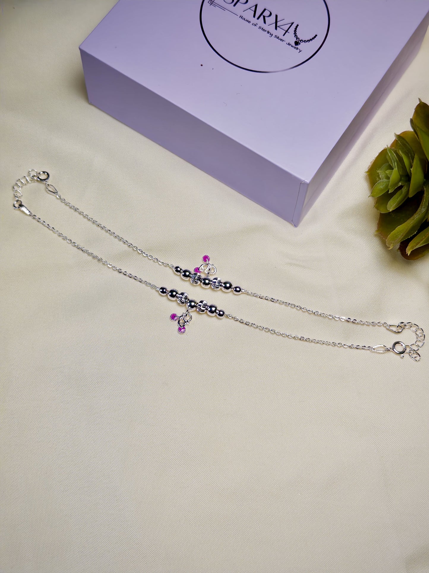 Kids silver anklets with purple accents on a light surface with a box in the background.