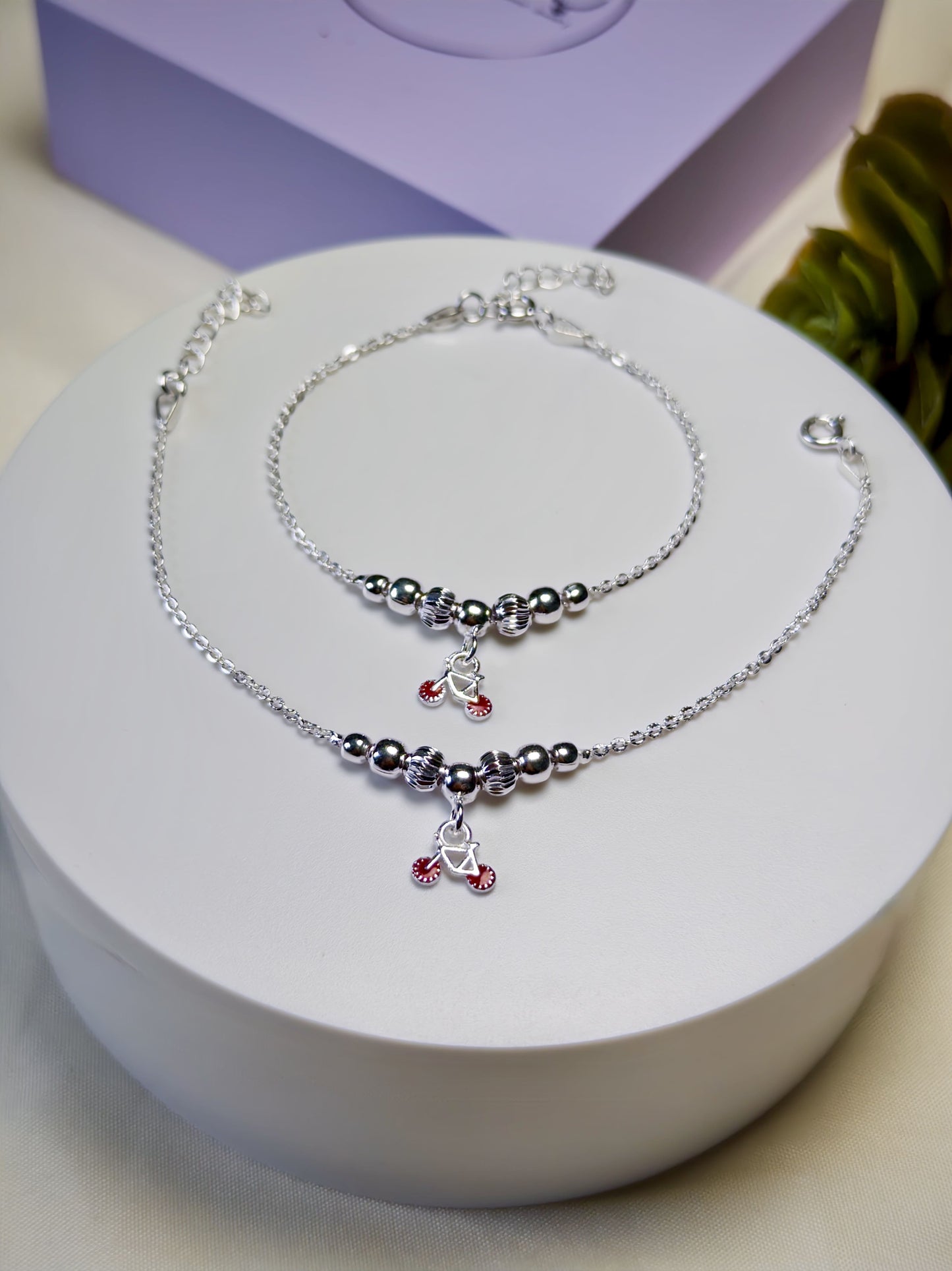 Kids silver anklets with red charms on a white stand with a purple box in the background