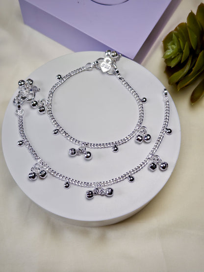 Pair of silver anklets on a white circular stand with a purple box and green leaves in the background.