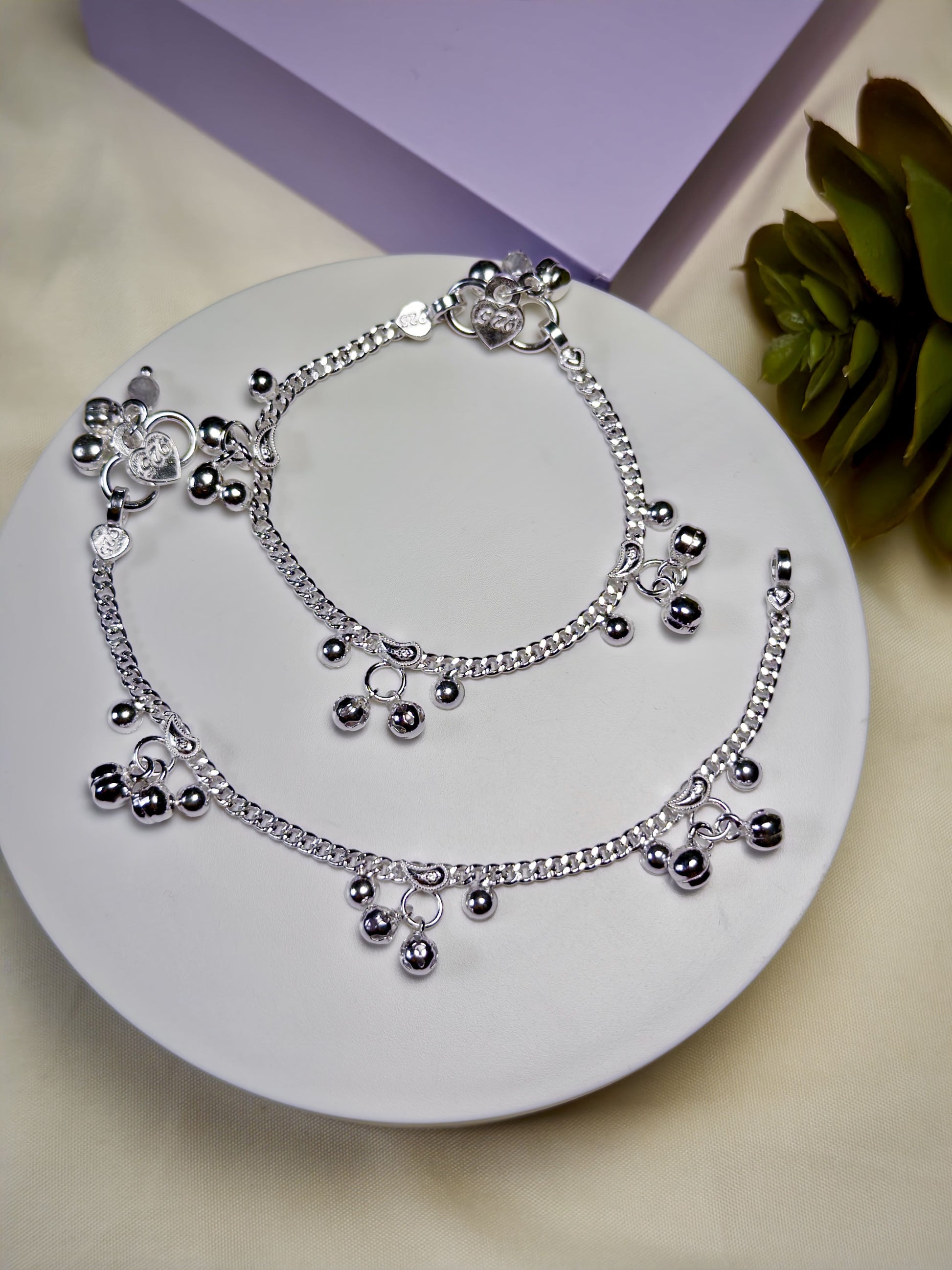 Kids silver anklets with charms on a white plate with a purple box and green leaves in the background