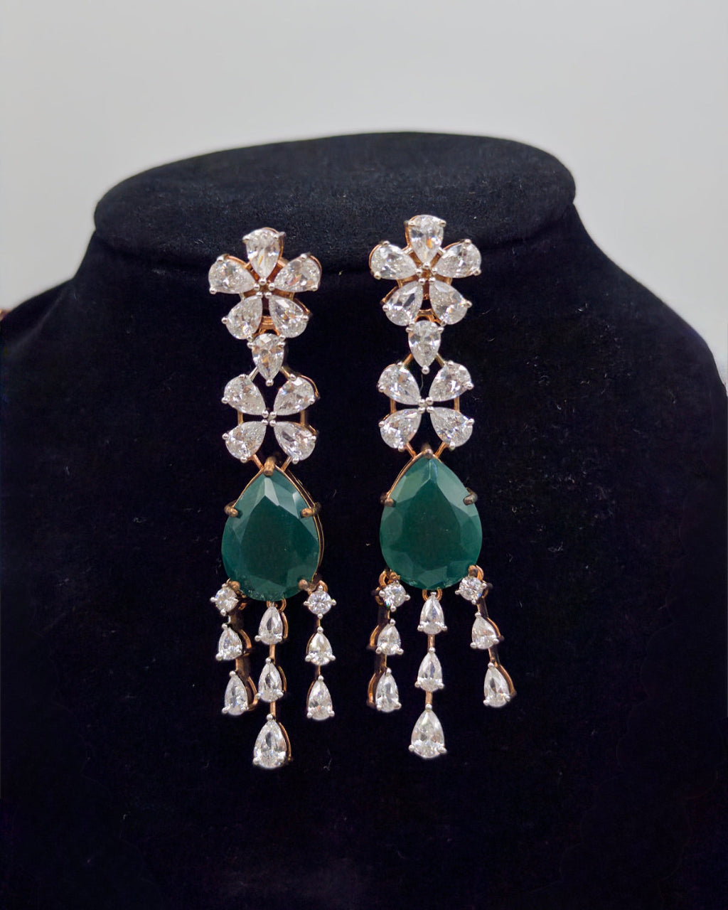 Pair of earrings with green teardrop and diamond-like stones on a black velvet stand.