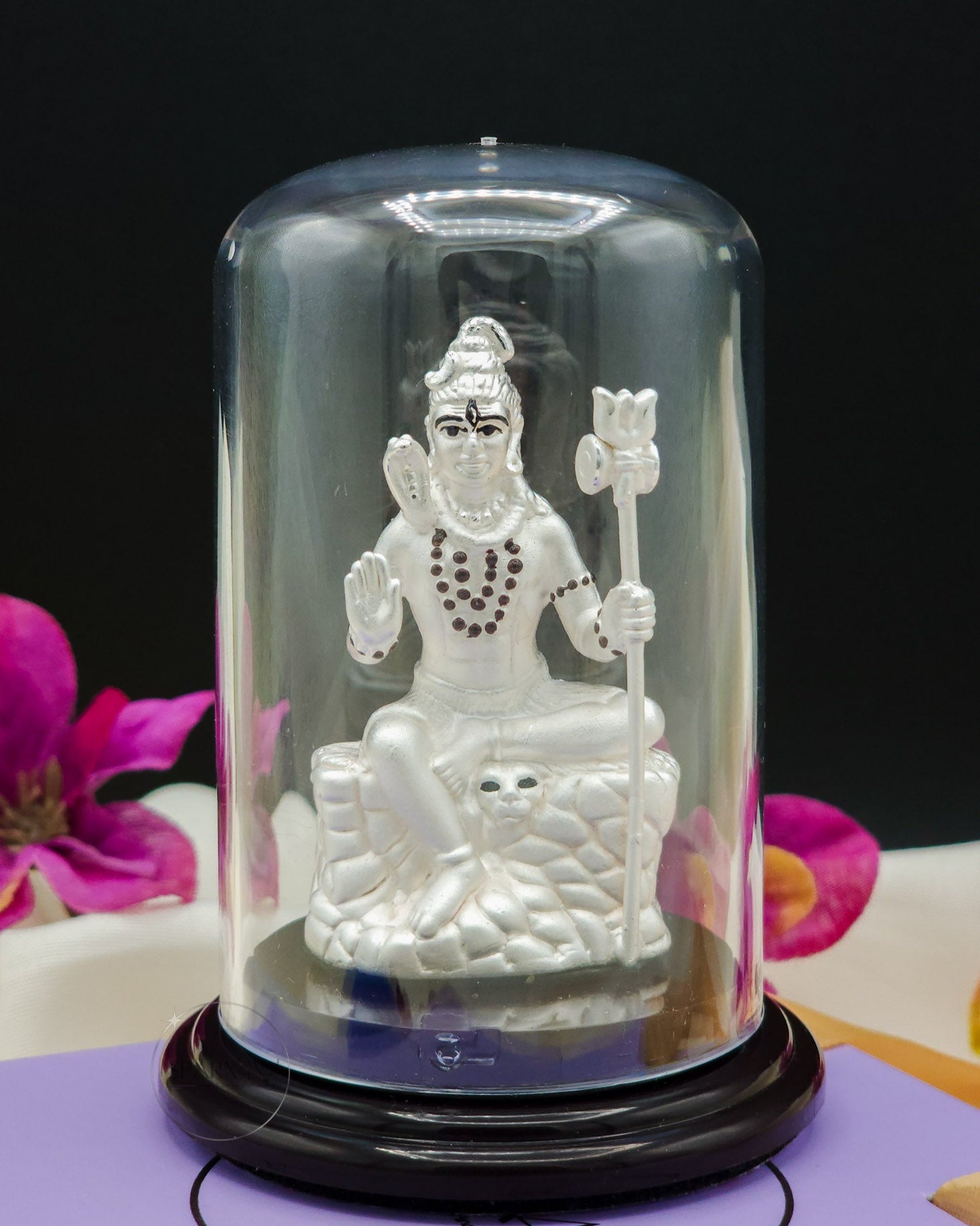 Silver statue of a Lord Shiva inside a glass dome with pink flowers in the background