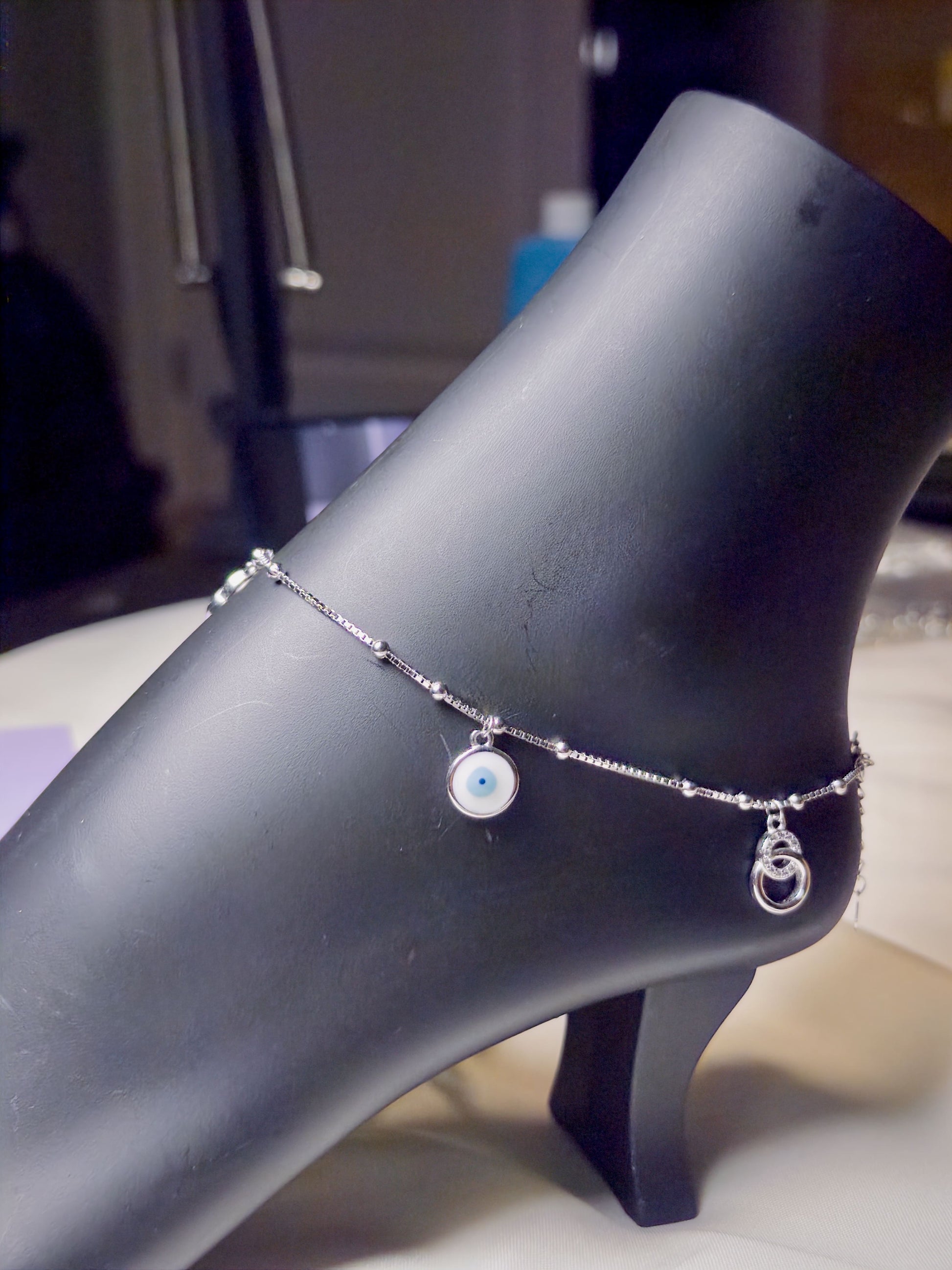 Silver anklet with charms on a black mannequin foot against a blurred indoor background