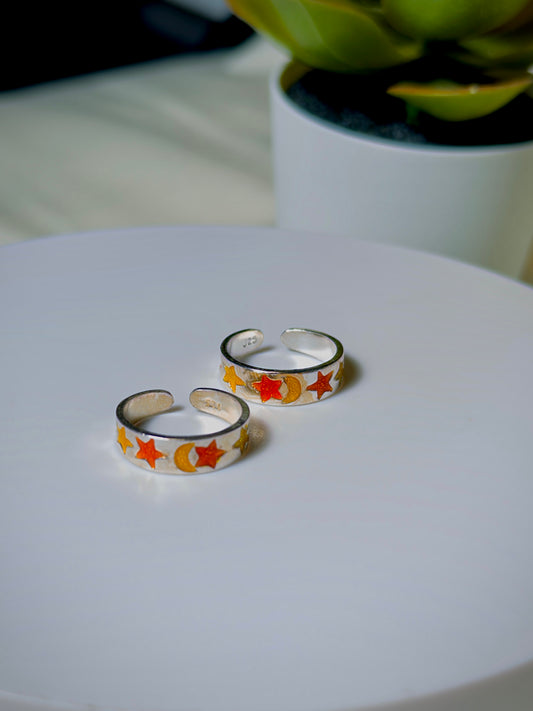 Silver toe rings with star and moon designs on a white surface, with a blurred plant in the background.