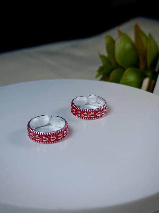 Two silver toe rings with red patterns on a white surface, with green leaves in the background.