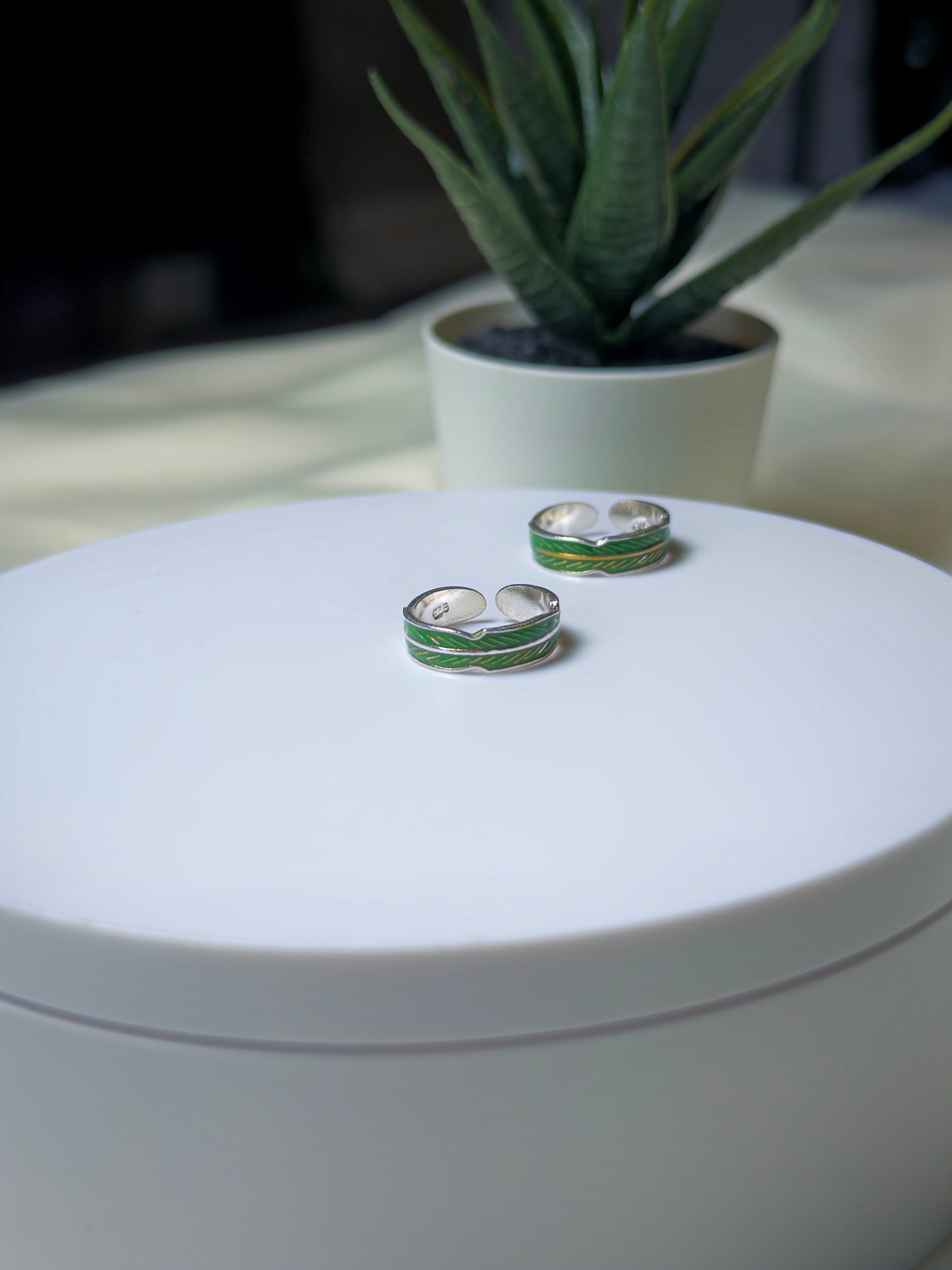 Silver toe rings with green accents on a white surface, with a plant in the background.