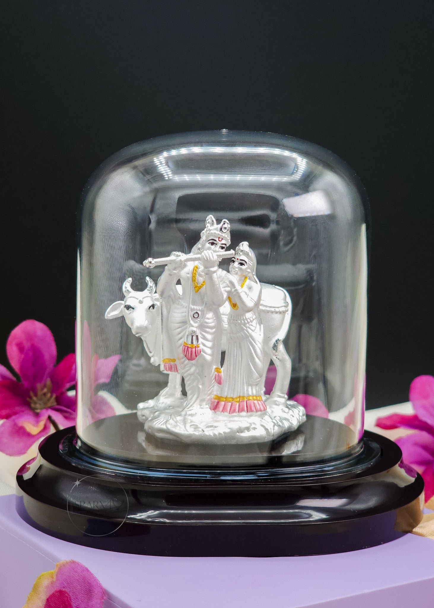 Silver Radha Krishna Idol in a glass dome with pink flowers in the background