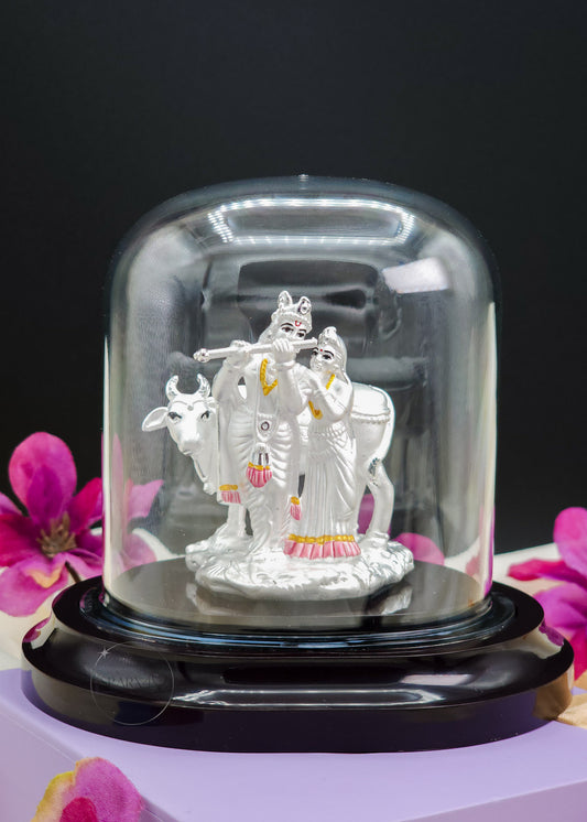 Silver Radha Krishna Idol in a glass dome with pink flowers in the background