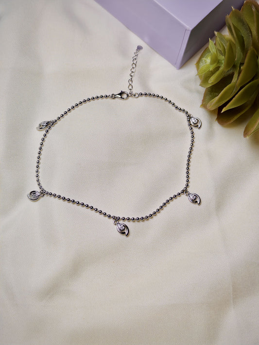 Silver bracelet with heart-shaped charms on a beige surface with a green plant and purple box in the background.