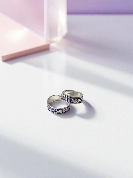 Two silver toe rings with black patterns on a light pink background