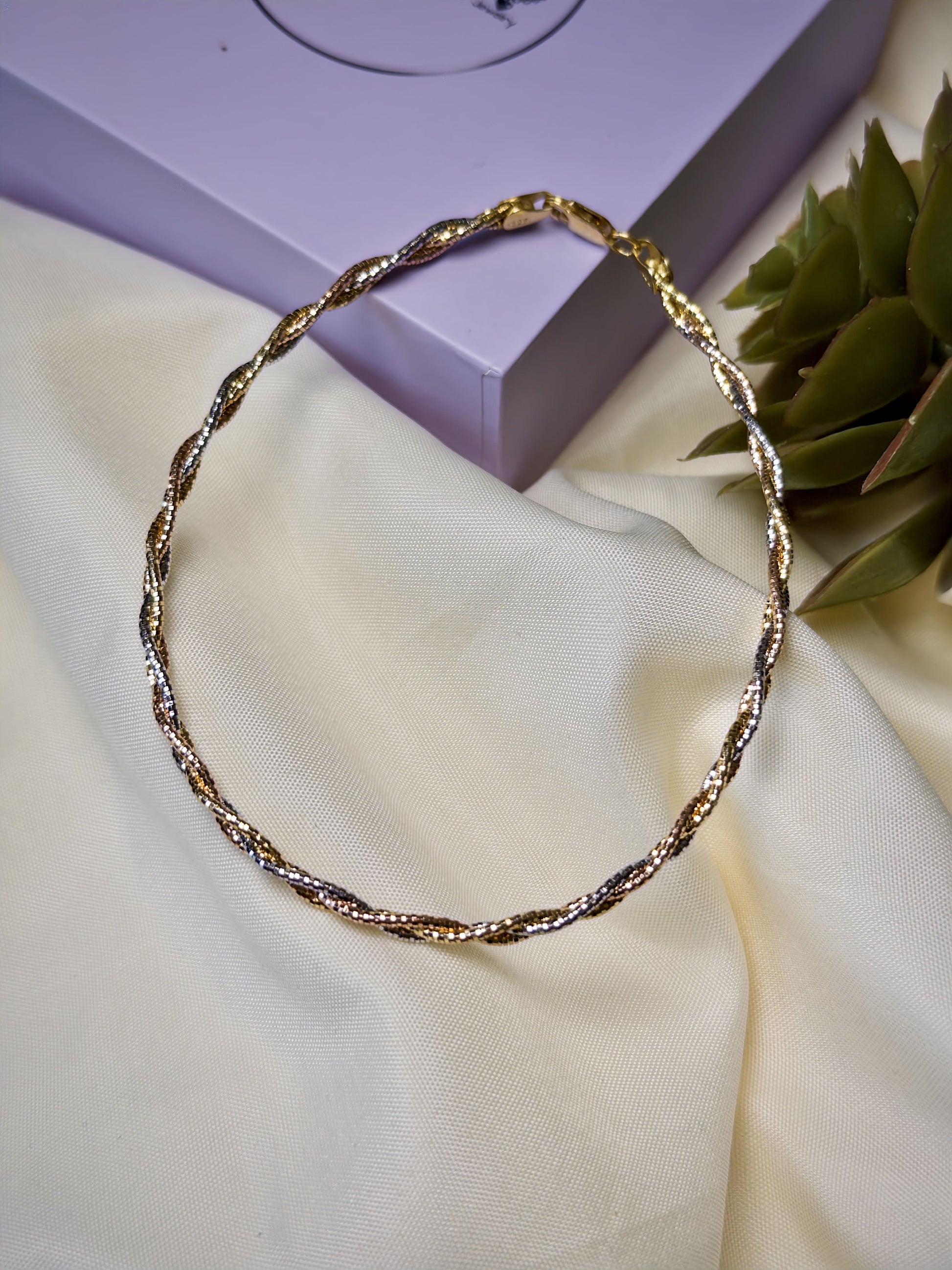 Twisted gold anklet on a light fabric surface with a purple box and green plant in the background