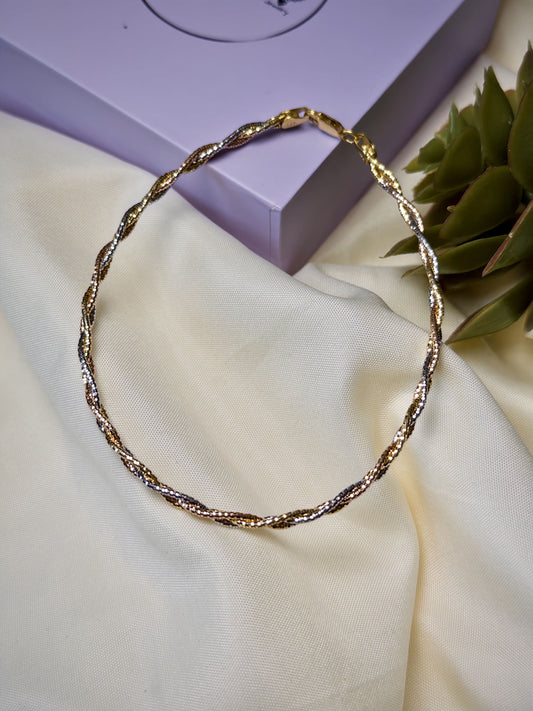 Twisted gold anklet on a light fabric surface with a purple box and green plant in the background
