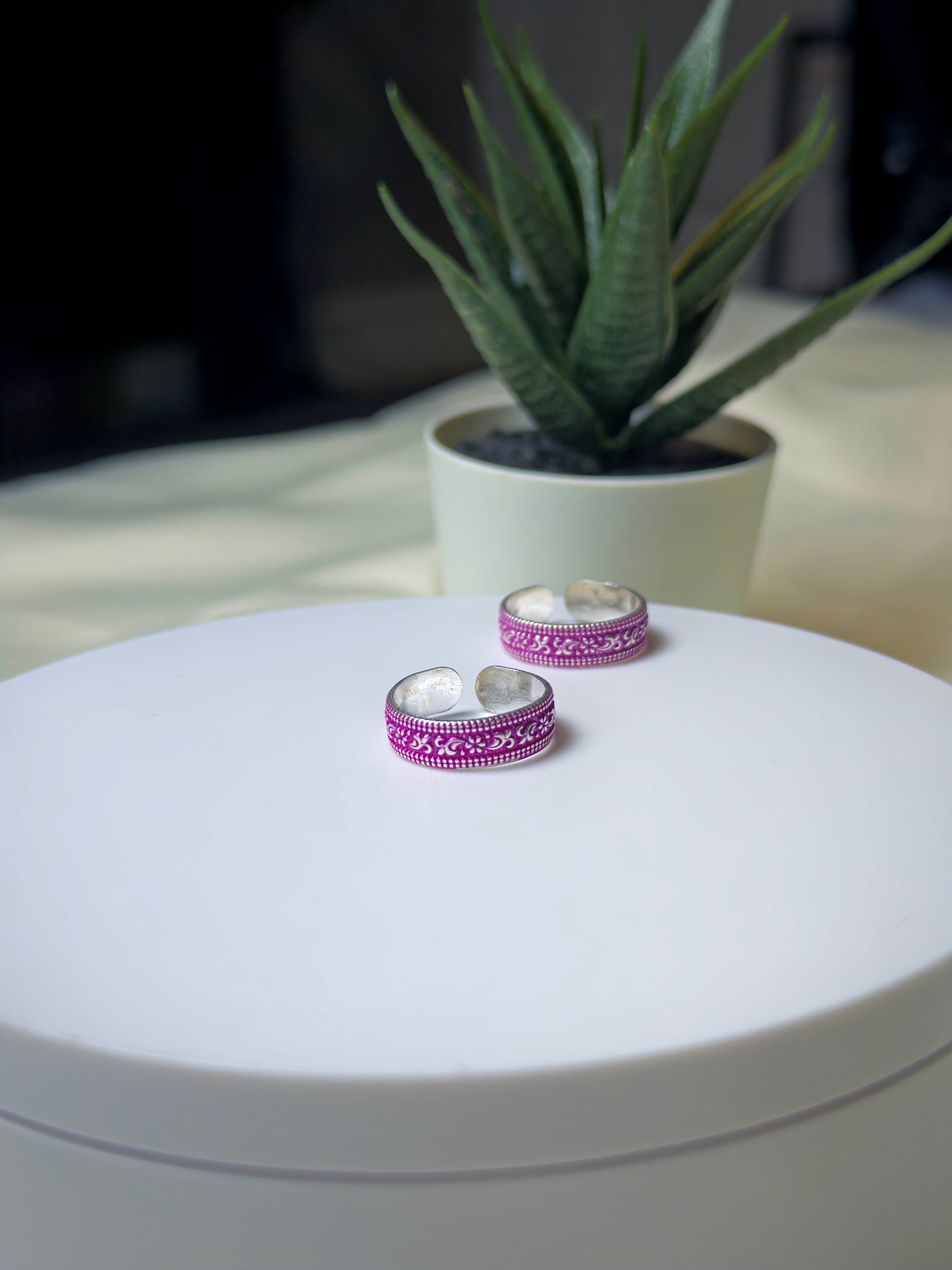 Two silver toe rings with pink patterns on a white surface, with a plant in the background.